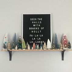 there are many small christmas trees on the shelf in front of this sign that says deck the hall's with boughs of holly fa la la la la la la la