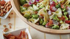 a wooden bowl filled with pasta salad next to two small bowls full of chopped nuts