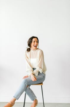 a woman sitting on top of a wooden chair in front of a white wall smiling