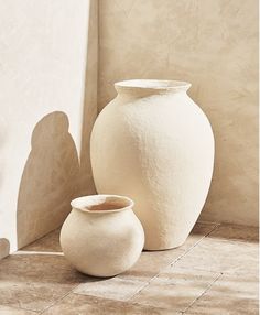 two white vases sitting next to each other on the floor in front of a wall