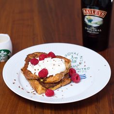 a white plate topped with french toast covered in whipped cream and raspberries next to a bottle of bailey's