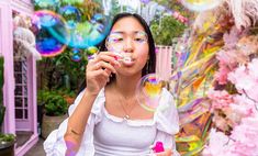 a young woman blowing bubbles in front of a pink building with lots of fake flowers