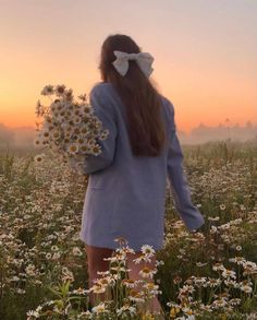 a woman standing in a field with daisies and looking at the sun setting behind her