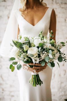 a bride holding a bouquet of white flowers and greenery in her hands with a brick wall behind her