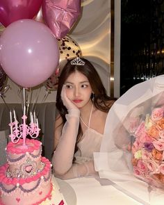 a woman sitting in front of a pink cake with candles on it and balloons behind her