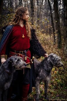 a man with long hair and beard standing next to two dogs in the woods,