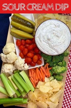 a wooden tray topped with veggies and dip next to crackers on a red checkered table cloth