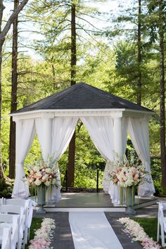 an outdoor wedding setup with white drapes and flowers on the aisle, surrounded by tall trees