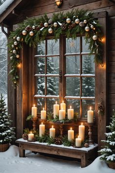 candles are lit in front of a window decorated with greenery and pine cones on a snowy day