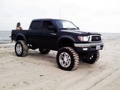 a woman sitting in the bed of a black truck on top of a sandy beach