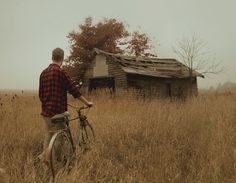 a man riding a bike through a field next to an old wooden house on a foggy day