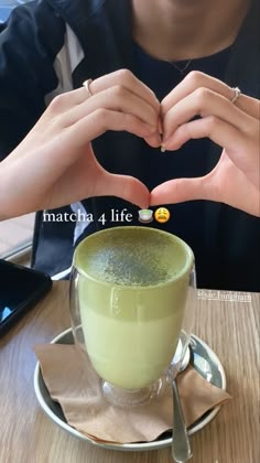 a person making a heart shape with their hands over a green liquid in a cup