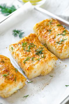 three fish fillets on a white plate with herbs and seasoning in the background