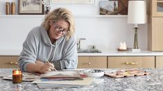 a woman sitting at a kitchen table writing in her notebook with candles on the counter