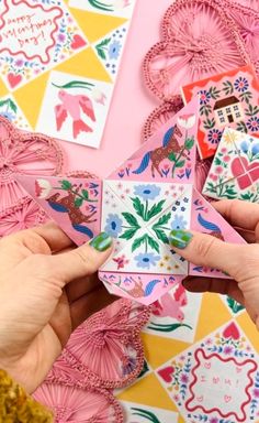 two hands are holding up some paper decorations on a pink background with other items in the background