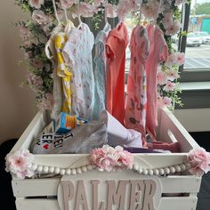 a white wooden box filled with pink and blue clothes next to a flower covered window