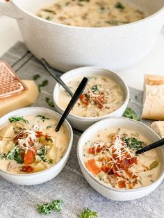 three white bowls filled with soup next to some bread and cheese on a tablecloth