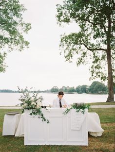 a person sitting at a table with flowers and greenery in front of it, surrounded by trees