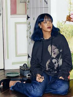 a woman with blue hair sitting on the floor wearing a black hoodie and jeans