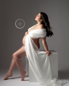 a pregnant woman in white is sitting on a stool and posing for the camera with her legs crossed