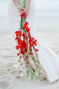 a woman in white dress holding red flowers on the beach with an angel behind her