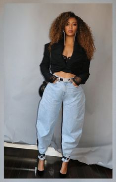 a woman standing in front of a gray wall wearing blue jeans and a black shirt