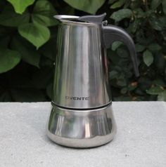 a stainless steel coffee pot sitting on top of a cement table next to green plants