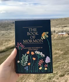 the book of mormon is held up in front of an open field with wildflowers