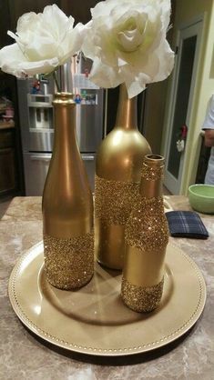 three gold vases with white flowers in them on a counter top next to a plate