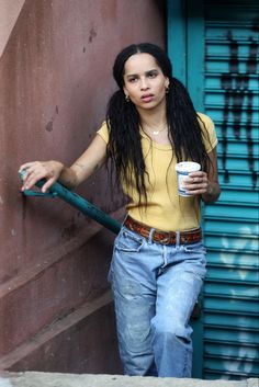 a woman with dreadlocks holding a coffee cup and leaning against a building wall