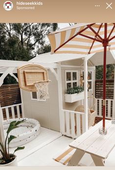 a small white house with an umbrella over it's door and deck area, next to a potted plant