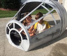 two children are sitting in a metal car shaped like a wheelbarrow with wheels