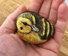 a hand holding a painted rock with a yellow and black bird on it's face