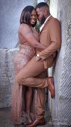 a man and woman leaning against a stone wall in formal wear, smiling at each other