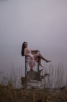 a woman is sitting in a chair by the water on a foggy day with her legs crossed