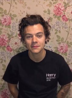 a young man with curly hair wearing a harry potter t - shirt in front of a floral wallpaper