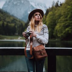 a woman standing on a bridge holding a camera in her hand and wearing a hat