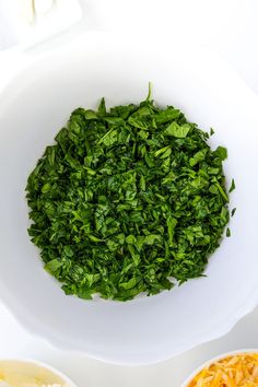 a white bowl filled with greens next to two bowls full of cheese and other foods