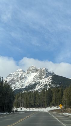 an empty road in front of a mountain range with snow on the top and trees below