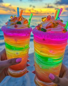 two people holding up colorful drinks with straws on the beach in front of an ocean