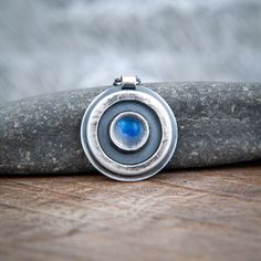 A lovely clear moonstone with deep blue fire.  Simply set in a disc pendant, and encircled with a raised halo. I have oxidized the silver and polished it to highlight the raised features.  The reverse of the pendant shows the hammer marks made by the forging process.   Stamped 925 on reverse for Sterling Silver.  A simple tube creates a bail for the chain - a fine oval link chain with bolt ring closure.  (If your preferred chain length is not in the list of options, just contact me). Simple, yet Blue Moonstone Necklaces With Moon Phase Detail, Blue Moonstone Necklaces With Moon Phase, Blue Moonstone Necklace With Moon Phase Detail, Blue Moonstone Round Jewelry, Blue Moonstone Pendant Jewelry, Blue Round Moonstone Jewelry, Blue Moon Phase Round Pendant Jewelry, Blue Round Labradorite Jewelry, Nickel Free Moonstone Necklaces