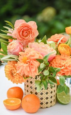 an arrangement of flowers and oranges on a white table with greenery in the background