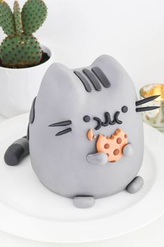 a grey cat cake sitting on top of a white plate next to a cacti