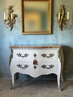 an ornate white dresser in front of a blue wall with a gold framed mirror on it