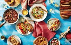 a table topped with plates of food next to hotdogs and other foods on paper plates