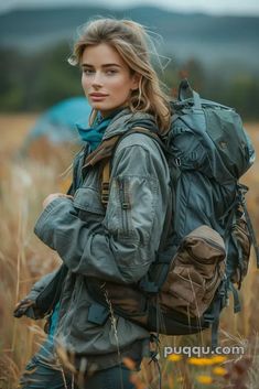 a woman with a backpack is standing in tall grass and looking at the camera while holding her hands behind her back