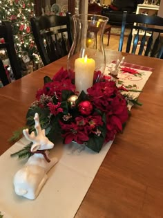 a christmas centerpiece with red flowers and greenery on a wooden table in front of a lit candle