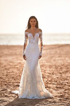 a woman standing on top of a sandy beach wearing a wedding dress with long sleeves