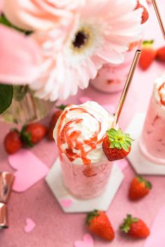 two glasses filled with ice cream and strawberries on a pink table next to flowers