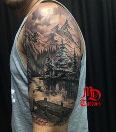 a man with a mountain scene tattoo on his arm and shoulder is standing in front of a body of water